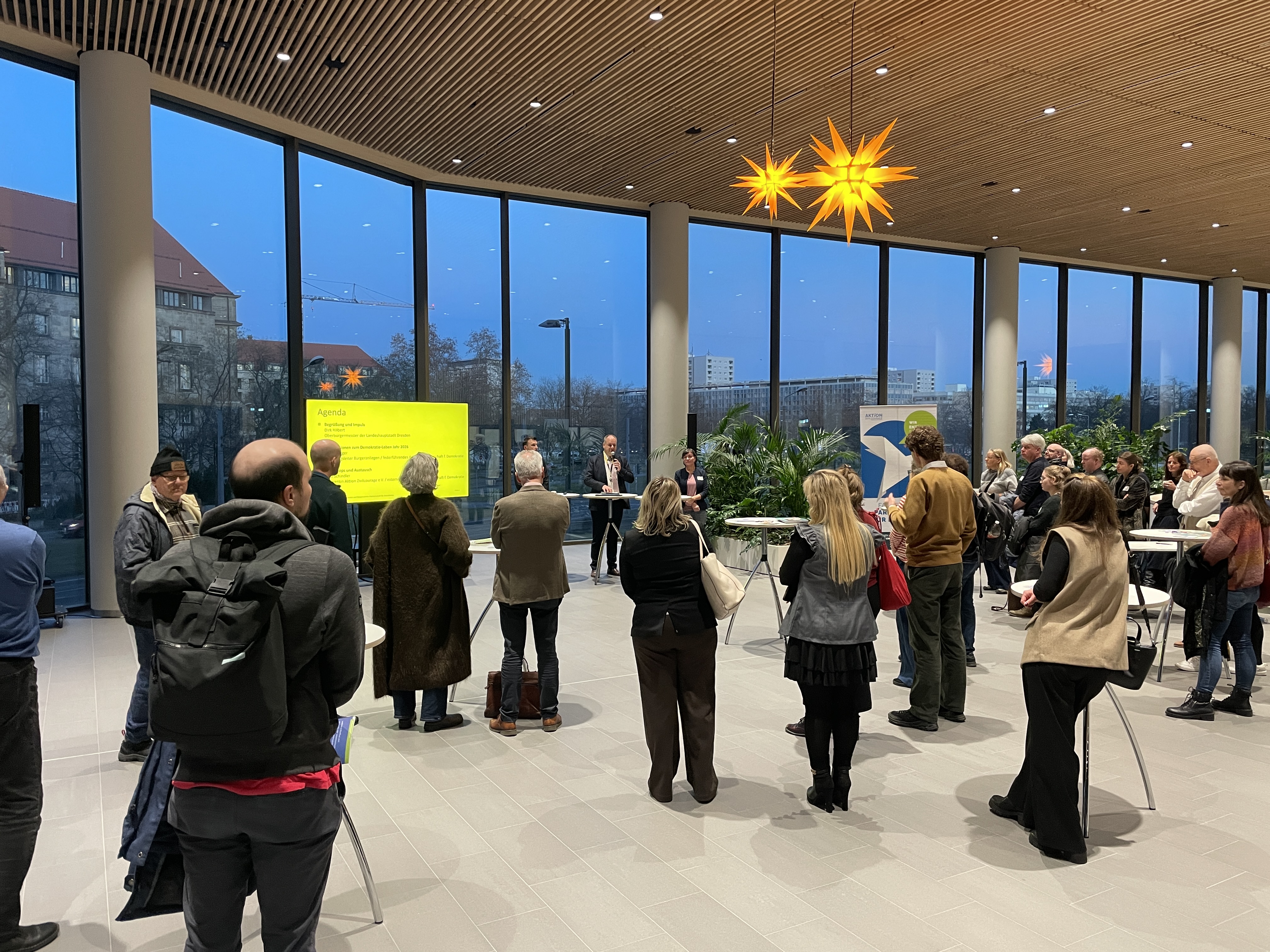 KI generiert: Das Bild zeigt eine Gruppe von Menschen, die sich in einem modernen Veranstaltungsraum versammelt haben, um einer Präsentation oder Rede zuzuhören. Im Hintergrund sind große Fenster und stimmungsvolle Beleuchtung zu sehen.